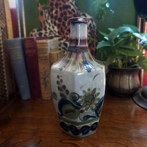 May include: A hexagonal ceramic bottle featuring a white base with hand-painted floral motifs in shades of blue, green, and brown. The bottle has a narrow neck with a metallic rim and a tapered body that widens toward the base with decorative patterns.