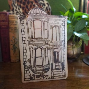 May include: A rectangular ceramic tile featuring a dark brown line drawing of a multi-story Victorian-style house with a bay window, a front door with steps, and a small tree at the base. The tile has a cream-colored background and rounded top.