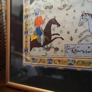 May include: A framed miniature painting depicting a polo match with riders on horseback. The scene features figures in orange and blue attire on dark brown and white horses, set against a light background with green grass. A decorative border surrounds the art.