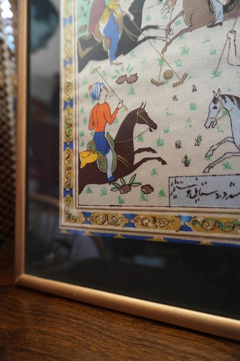 May include: A framed miniature painting depicting a polo match with riders on horseback. The scene features figures in orange and blue attire on dark brown and white horses, set against a light background with green grass. A decorative border surrounds the art.