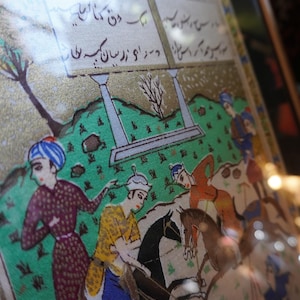 May include: A framed miniature painting depicting a polo match on a green field with figures on horseback. The artwork features intricate gold detailing, vibrant colors, and Persian script written in black ink within rectangular panels at the top.