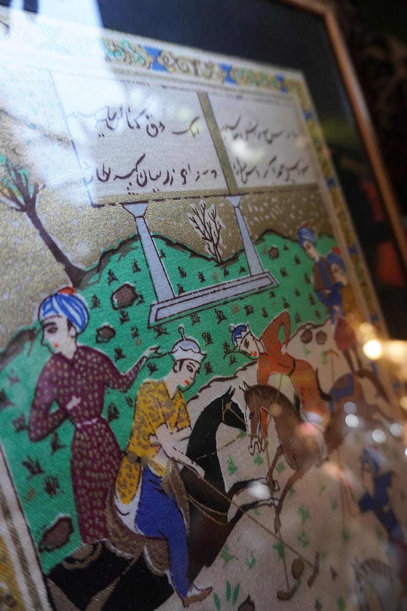 May include: A framed miniature painting depicting a polo match on a green field with figures on horseback. The artwork features intricate gold detailing, vibrant colors, and Persian script written in black ink within rectangular panels at the top.