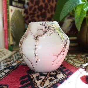 Puede incluir: Un pequeño jarrón de cerámica blanquecina con un diseño orgánico único. El jarrón presenta una red de líneas marrones oscuras en su superficie. El jarrón está sobre una alfombra estampada, con libros y una planta al fondo.