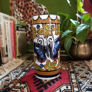 May include: A cylindrical ceramic vase with a butterfly design. The vase features a white background with a large blue butterfly, surrounded by green and yellow floral accents. The top and bottom have brown and blue bands.