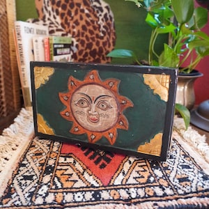 Könnte beinhalten: Eine rechteckige Holzkiste mit einem handgemalten Sonnendesign. Die Sonne hat ein lächelndes Gesicht und ist von orangefarbenen Strahlen umgeben. Die Kiste ist grün mit goldenen Eckakzenten und liegt auf einem gemusterten Teppich.