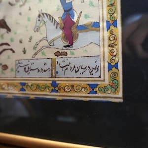 May include: A framed miniature painting depicting a figure in a blue tunic and purple trousers riding a white horse. The artwork features intricate gold and blue decorative borders and includes black Persian script text at the bottom of the scene.