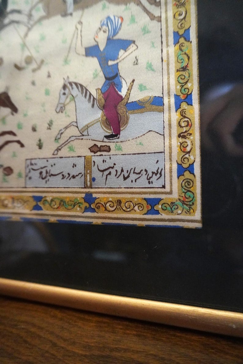 May include: A framed miniature painting depicting a figure in a blue tunic and purple trousers riding a white horse. The artwork features intricate gold and blue decorative borders and includes black Persian script text at the bottom of the scene.