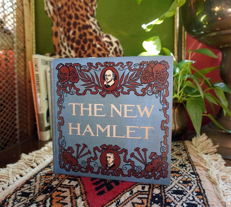 May include: A hardcover book titled "The New Hamlet" with a light blue cover. The cover features decorative red and brown accents, including portraits and theatrical masks. The book is displayed on a patterned rug.