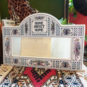 Pode incluir: Moldura de foto de cerâmica branca com um design floral e as palavras "Home Sweet Home" em letras pretas. A moldura tem um inserto de vidro transparente.