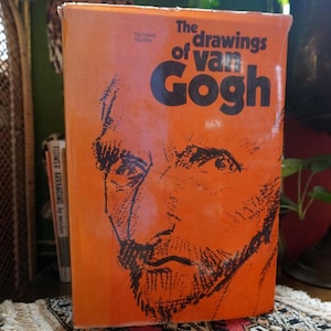 Puede incluir: Un libro naranja titulado "The Drawings of Van Gogh" con una ilustración negra del rostro de Van Gogh. El libro está de pie sobre una alfombra estampada, con otros libros y plantas en el fondo.