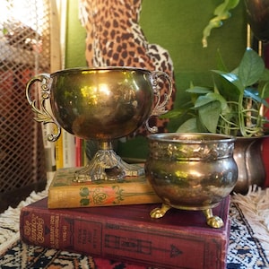 Może przedstawiać: Dwie antyczne mosiężne misy stoją na starych książkach. Większa misa ma ozdobne uchwyty i podstawę na cokole. Mniejsza misa spoczywa na trzech łapach. Książki noszą tytuły "Junior Classics" i "Folk Tales and Myths".