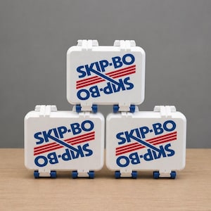 May include: Three white SKIP-BO game boxes stacked on a wooden surface. Each box has the game's logo in blue and red. The boxes have blue wheels and white latches.