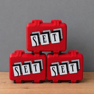 May include: Three red miniature suitcases stacked, each with the word "SET" in white block letters on a black and white background. The suitcases have black hinges and latches.