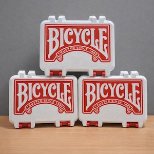 May include: Three white Bicycle card cases with red accents and the brand name in red. Each case has the text "TRUSTED SINCE 1885". The cases are stacked on a wooden surface.