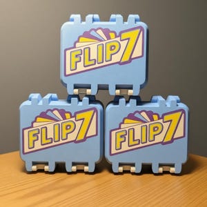 May include: Three light blue toy cases stacked. Each case has the text "FLIP 7" in yellow and purple. The cases are on a wooden surface with a gray background.