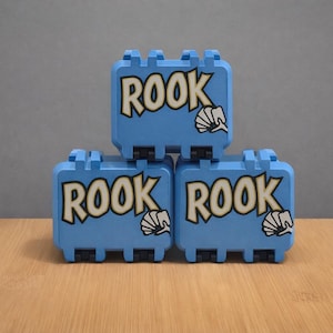 May include: Three light blue boxes stacked, each with the word "ROOK" in a bold, stylized font. A white graphic resembling a seashell is next to the word. The boxes have a rectangular shape with a handle and a locking mechanism.