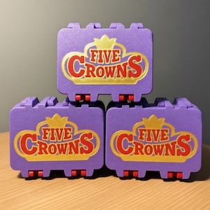 May include: Three purple Five Crowns game cases stacked on a wooden surface. Each case has a gold crown logo with the words "FIVE CROWNS" in red. The cases have red wheels.