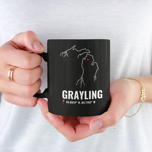 以下が含まれることがあります： 前面にミシガン州の白抜き輪郭と「Grayling 44.6614° N, 84.7148° W」の文字が印刷された黒いコーヒーカップ