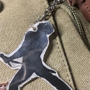 Op de afbeelding: Een zilveren sleutelhanger met een transparante plastic hanger met een zwart silhouet van een persoon die gitaar speelt. De sleutelhanger is bevestigd aan een bruine leren tas.