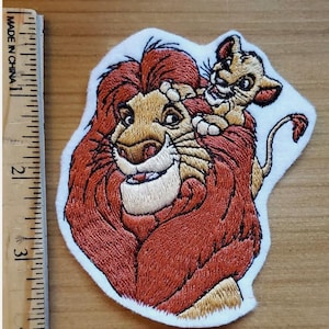 May include: Embroidered patch featuring a lion and cub from The Lion King. The lion has a red mane and a tan face, with the cub perched on its head. The patch is on a white background and has the text "MADE IN CHINA" on the side.