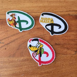 May include: Three Disney-themed patches. One features Tigger, another has "Hakuna Matata" text, and the third depicts Pluto. Each patch is in the shape of the Disney logo, with colorful character designs and text.