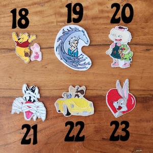 May include: A collection of embroidered patches featuring cartoon characters. Includes Winnie the Pooh, Elsa from Frozen, a Rugrats character, a mummy cat, a dog in a yellow car, and Bugs Bunny.