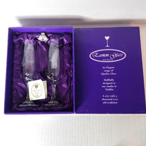 May include: A set of two etched champagne flutes in a purple box. The flutes feature bride and groom designs. The box has the text "Eamon Glass Dublin" and "An Elegant range of Quality Glass".