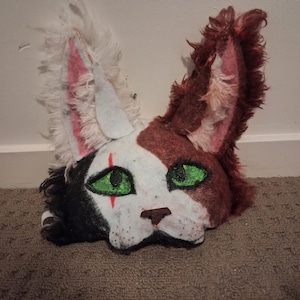 May include: A handcrafted cat mask featuring a split-color design with white, black, and reddish-brown faux fur. The mask has large pointed ears, bright green eyes with a red vertical line under the left eye, a small brown nose, and white whiskers.