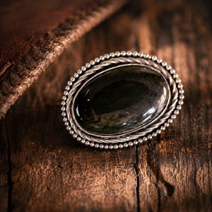 May include: An oval brooch featuring a dark green, polished stone cabochon set in a silver-toned metal frame. The setting is detailed with a twisted rope-like inner border and an outer edge composed of small, uniform metallic beads.