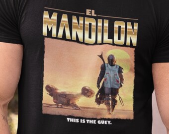 Può includere: T-shirt nera con una grafica dorata di un personaggio della serie Star Wars, The Mandalorian. La grafica include il testo "El Mandalon" e "This is the guy."