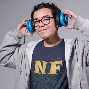 May include: A navy blue unisex softstyle t-shirt with the letters "NF" in gold. The shirt has a matrix-style design and size chart in inches. The model is wearing headphones and glasses.