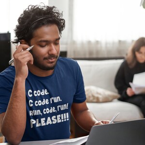 May include: A navy blue t-shirt with the text "C code. C code run. Run, code, run! PLEASE!!!" in white. A person is wearing the shirt and holding a pen, looking at a laptop. Another person is in the background.