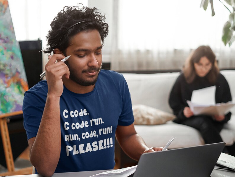 May include: A navy blue t-shirt with the text "C code. C code run. Run, code, run! PLEASE!!!" in white. A person is wearing the shirt and holding a pen, looking at a laptop. Another person is in the background.