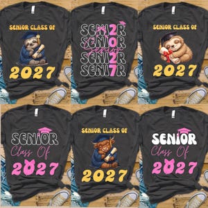 Può includere: Una collezione di sei magliette grigio scuro con vari design grafici per la classe di diploma del 2027. Le stampe includono illustrazioni di un bradipo, una mucca highland e una tipografia stilizzata nei colori rosa, bianco e giallo.
