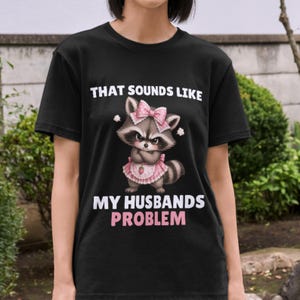 Pode incluir: Camiseta preta com um guaxinim de desenho animado com laço e vestido rosa, e o texto "THAT SOUNDS LIKE MY HUSBANDS PROBLEM" em branco e rosa. O guaxinim tem uma expressão severa. A camiseta é de gola redonda.