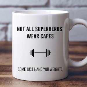 May include: White ceramic mug with black text that reads "Not all superheroes wear capes. Some just hand you weights." A black dumbbell icon is between the two lines of text.