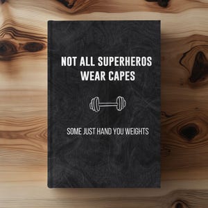 May include: A black notebook with the text "NOT ALL SUPERHEROES WEAR CAPES" and "SOME JUST HAND YOU WEIGHTS" in white. A dumbbell graphic is between the two phrases. The notebook is on a wooden surface.