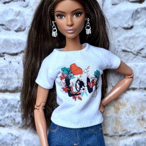 May include: A doll wearing a white t-shirt with a floral and text design that says "LOVE", and blue denim jeans. The doll has long brown hair, silver earrings, and is posed against a textured, light-colored background.