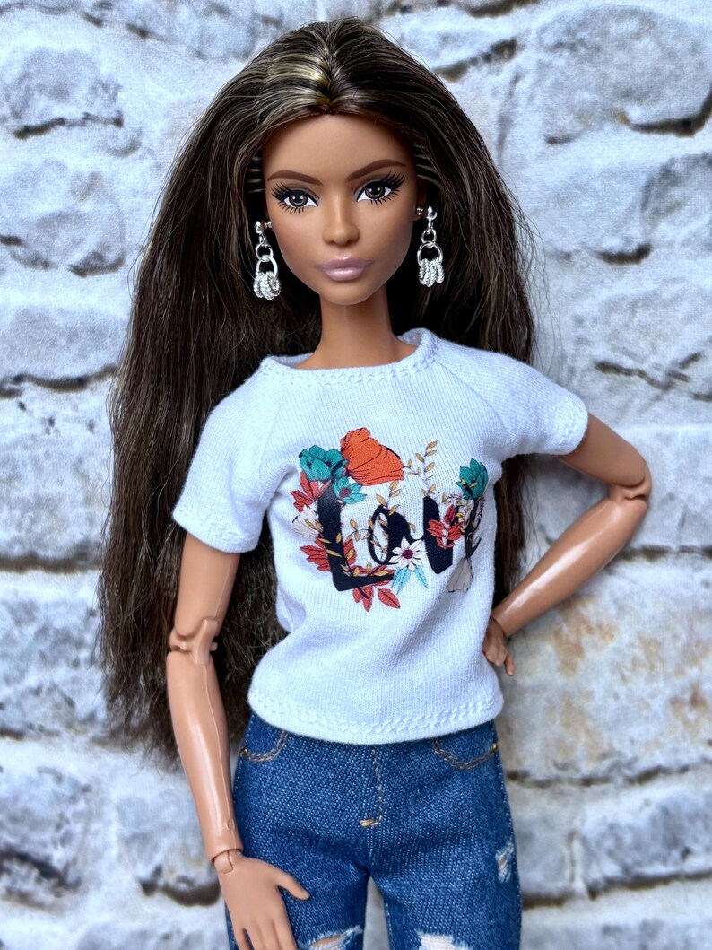 May include: A doll wearing a white t-shirt with a floral and text design that says "LOVE", and blue denim jeans. The doll has long brown hair, silver earrings, and is posed against a textured, light-colored background.