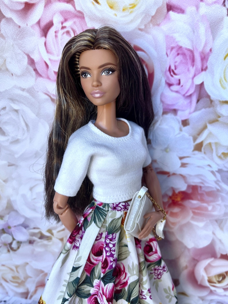 Peut inclure: Poup&eacute;e mannequin aux longs cheveux bruns, v&ecirc;tue d'un haut court blanc et d'une jupe &agrave; imprim&eacute; floral avec des fleurs violettes et roses. La poup&eacute;e tient un sac &agrave; main blanc avec une cha&icirc;ne dor&eacute;e. Le fond pr&eacute;sente un motif floral.