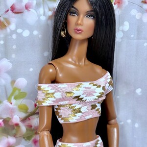 May include: A fashion doll with long black hair and brown eyes, wearing a two-piece outfit. The off-the-shoulder crop top and skirt feature a pink, white, and gold geometric pattern. The doll has gold hoop earrings.