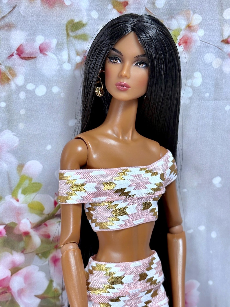 May include: A fashion doll with long black hair and brown eyes, wearing a two-piece outfit. The off-the-shoulder crop top and skirt feature a pink, white, and gold geometric pattern. The doll has gold hoop earrings.