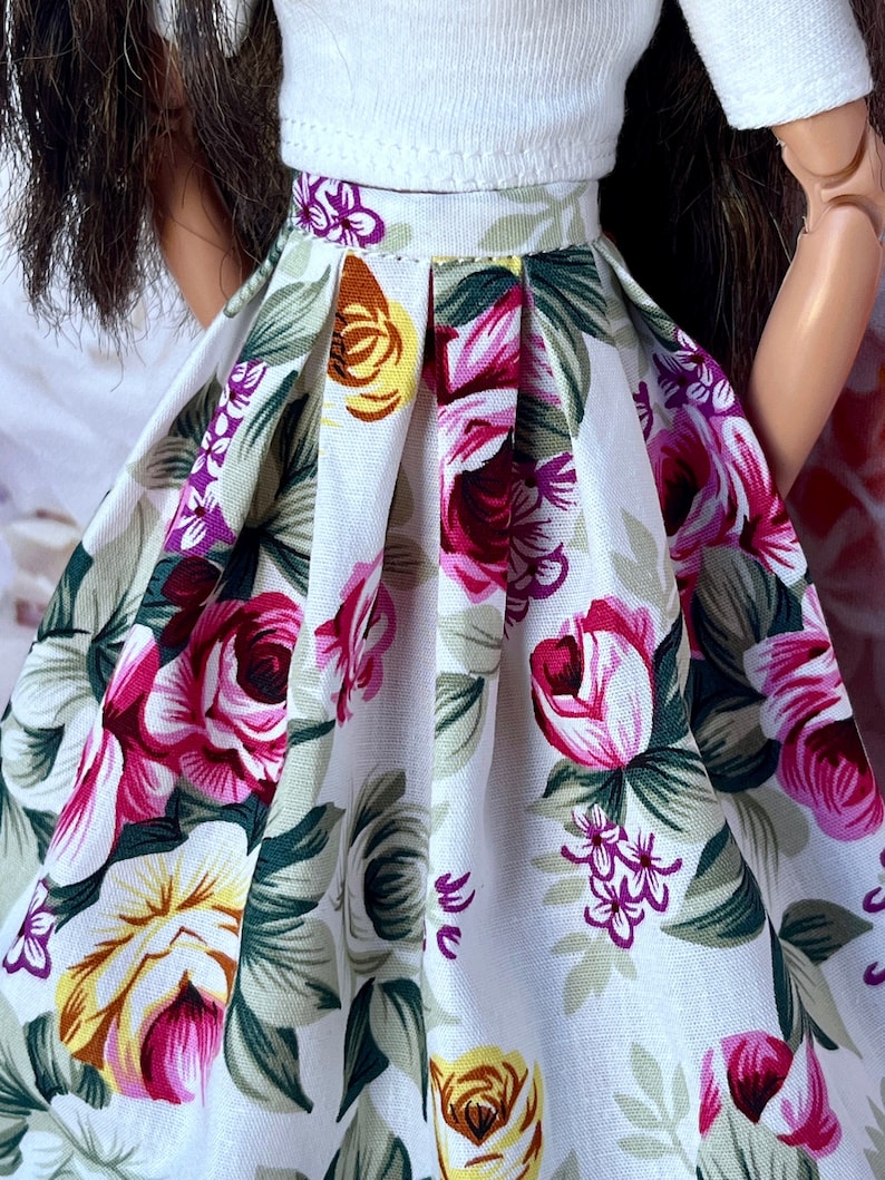 Peut inclure: Une poup&eacute;e porte un haut blanc et une jupe &agrave; fleurs. La jupe pr&eacute;sente un fond blanc avec de grandes roses roses, jaunes et violettes, ainsi que des feuilles vertes. La jupe est pliss&eacute;e &agrave; la taille.