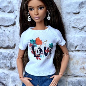 May include: A doll wearing a white t-shirt with a floral "Love" graphic and distressed blue jeans. The doll has long brown hair, silver earrings, and is posed against a textured background.