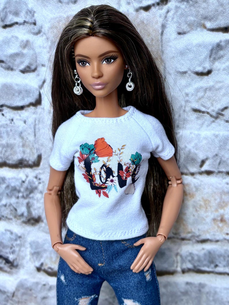 May include: A doll wearing a white t-shirt with a floral "Love" graphic and distressed blue jeans. The doll has long brown hair, silver earrings, and is posed against a textured background.