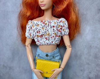 May include: A doll with long red hair wearing a white floral off-the-shoulder top, light blue jeans, and a yellow handbag.