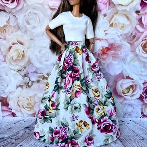 Peut inclure: Une poup&eacute;e v&ecirc;tue d'un haut blanc &agrave; manches courtes et d'une longue jupe fleurie. La jupe pr&eacute;sente un fond blanc avec de grandes roses color&eacute;es dans les tons de rose, de violet et de jaune, avec des feuilles vertes. La poup&eacute;e a de longs cheveux bruns.