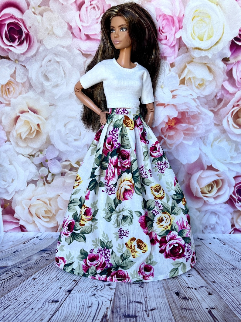 Peut inclure: Une poup&eacute;e v&ecirc;tue d'un haut blanc &agrave; manches courtes et d'une longue jupe fleurie. La jupe pr&eacute;sente un fond blanc avec de grandes roses color&eacute;es dans les tons de rose, de violet et de jaune, avec des feuilles vertes. La poup&eacute;e a de longs cheveux bruns.