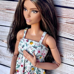 May include: A fashion doll with long brown hair and a floral print dress. The dress is white with a pattern of red, pink, and yellow flowers and green leaves. The doll is posed with one hand on its chest.