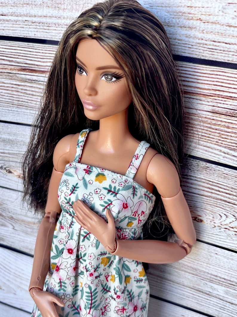 May include: A fashion doll with long brown hair and a floral print dress. The dress is white with a pattern of red, pink, and yellow flowers and green leaves. The doll is posed with one hand on its chest.